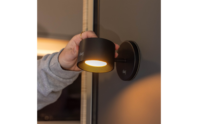 Hand mit Ring und grauem Ärmel hält schwarze zylindrische Wandlampe, die gelbes Licht abstrahlt; Lampenfuß an dunkler Wand montiert