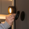 Hand mit Ring und grauem Ärmel hält eine runde schwarze Leuchte mit warmem Licht neben zwei runden schwarzen Scheiben an einer dunklen Wand