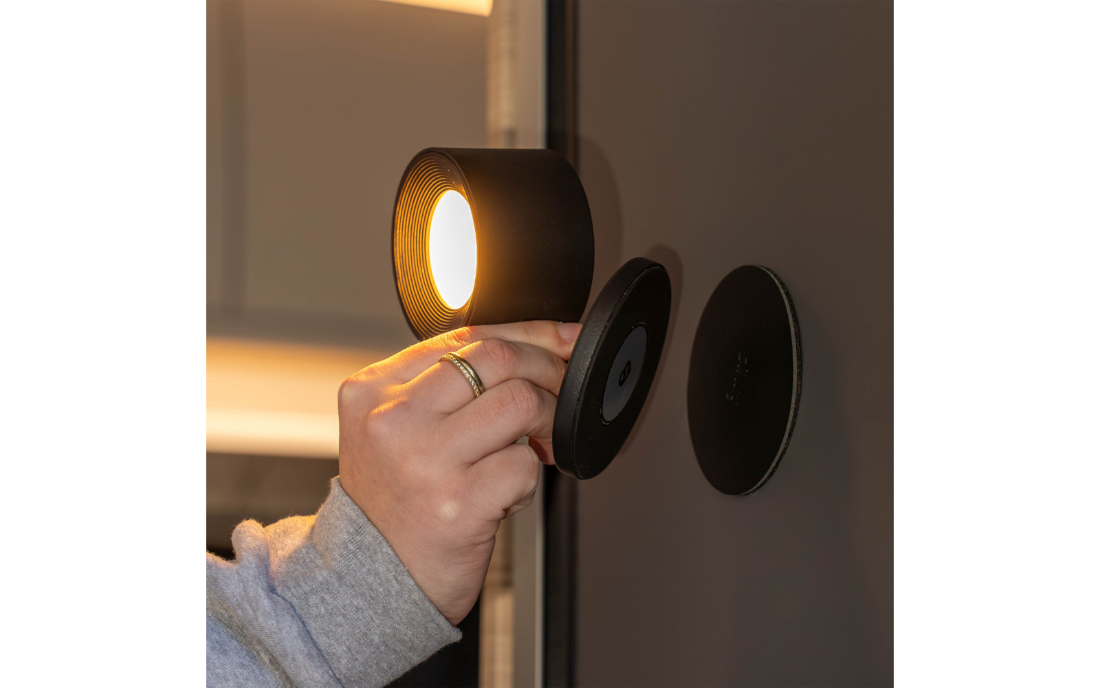 Hand mit Ring und grauem Ärmel hält eine runde schwarze Leuchte mit warmem Licht neben zwei runden schwarzen Scheiben an einer dunklen Wand
