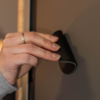 Hand mit Ring hält ein gebogenes schwarzes Gummiteil an einer grauen Fläche; Holzrahmen am Rand