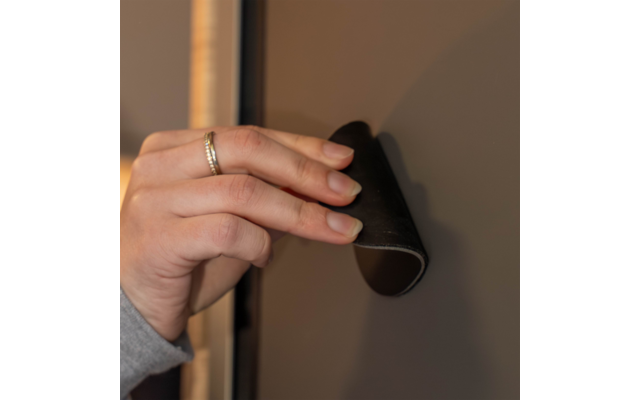 Hand mit Ring hält ein gebogenes schwarzes Gummiteil an einer grauen Fläche; Holzrahmen am Rand