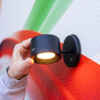 Hand an schwarzer zylindrischer Wandlampe mit runder Montageplatte, Lampe leuchtet, strukturierte Wand mit roten und grünen Flächen, Hand mit hellem Ärmel und Fingerringen