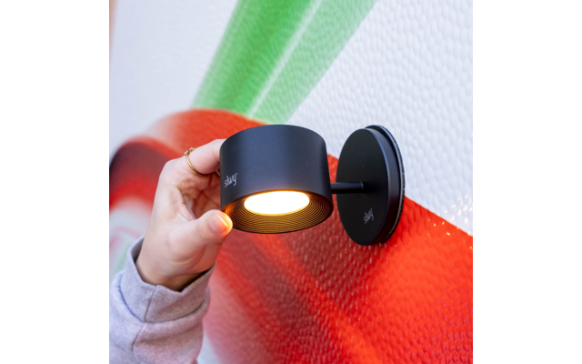 Hand an schwarzer zylindrischer Wandlampe mit runder Montageplatte, Lampe leuchtet, strukturierte Wand mit roten und grünen Flächen, Hand mit hellem Ärmel und Fingerringen