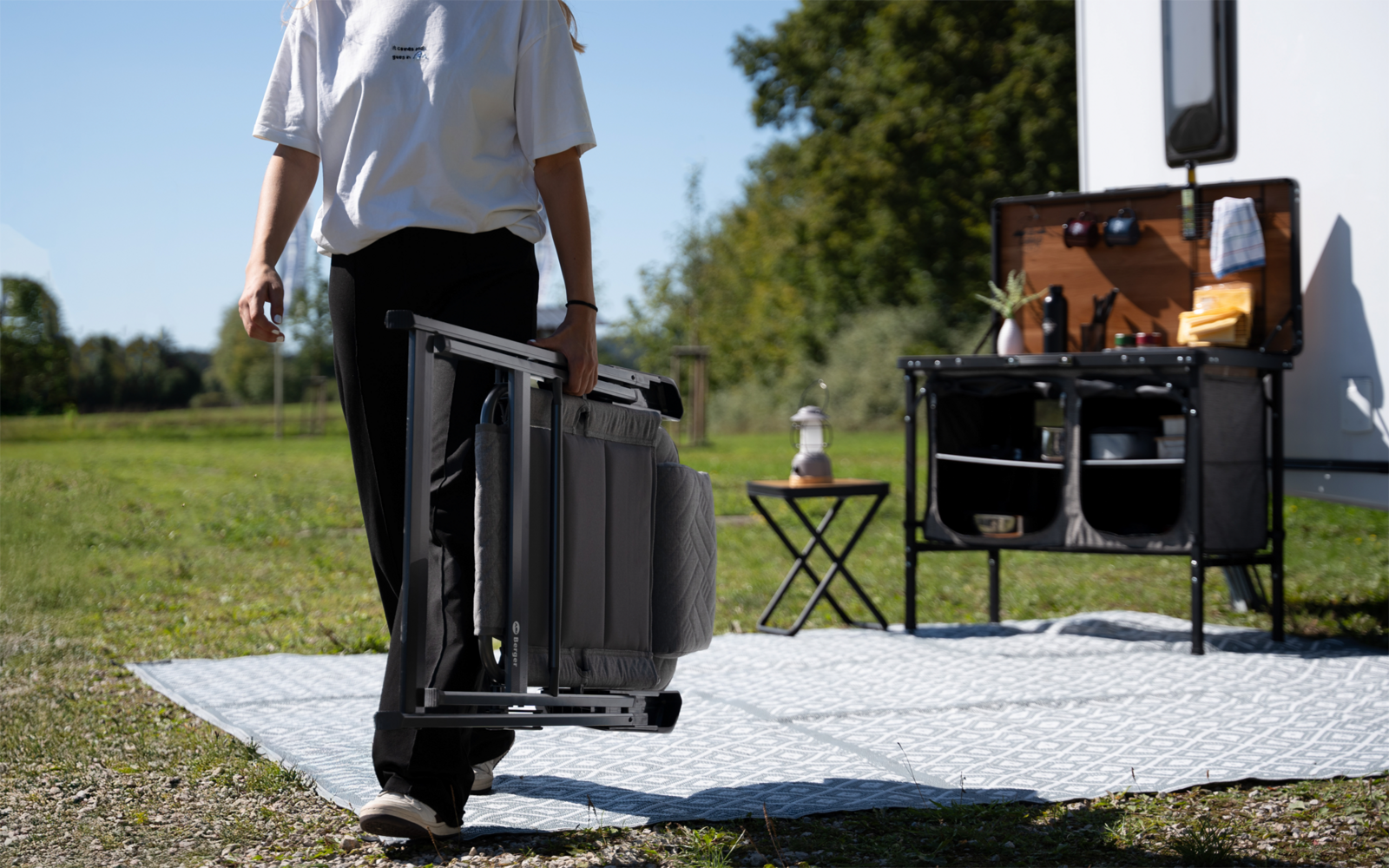 Person von hinten trägt einen zusammengeklappten grauen Campingstuhl; rechts ein Wohnwagen mit einer tragbaren Außenküche auf Beinen und offenen Fächern, darauf Flaschen und Tassen; kleiner Klapptisch mit Flasche auf einer hellen Bodenmatte, Kies, Wi