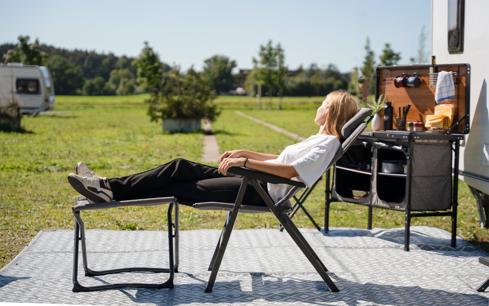 Person mit blondem Haar sitzt zurückgelehnt in einem Liegestuhl mit Fußstütze neben einem geparkten Wohnwagen und einer offenen Camping-Küchenbox; Wiese und Bäume im Hintergrund