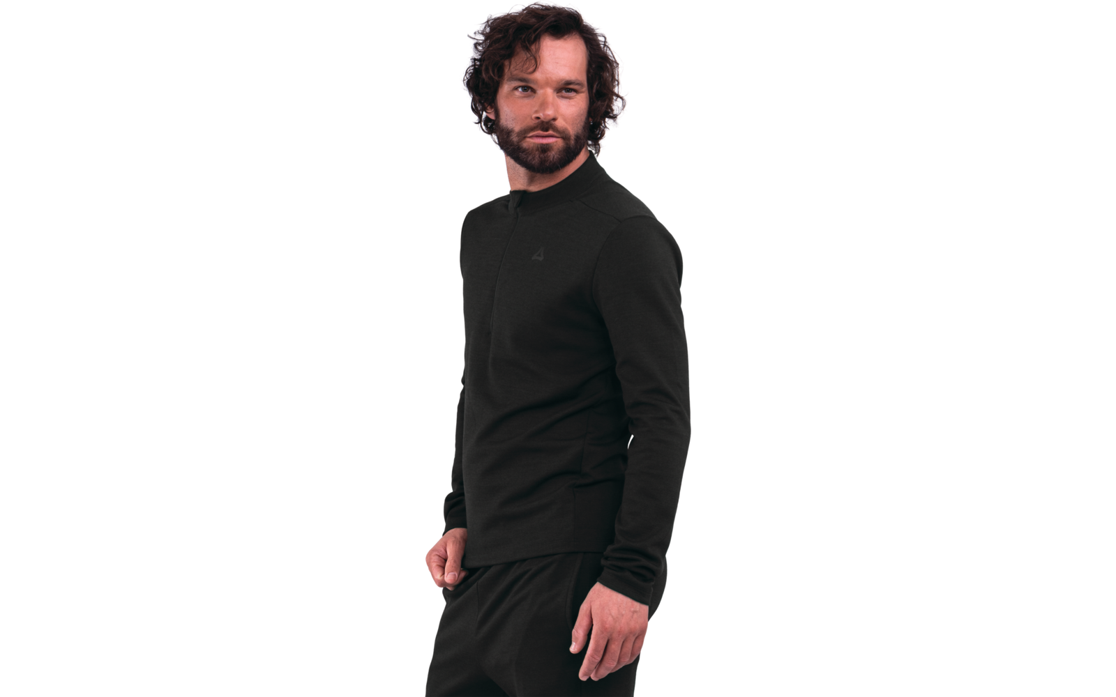 Persona di sesso maschile con capelli scuri e ricci e barba folta, indossa una maglia nera a maniche lunghe e pantaloni neri, in piedi davanti a uno sfondo bianco, leggermente girata di lato, mano sinistra parzialmente in tasca