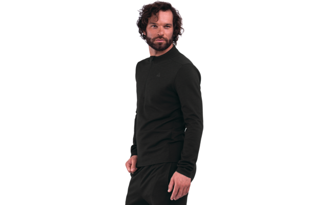 Persona di sesso maschile con capelli scuri e ricci e barba folta, indossa una maglia nera a maniche lunghe e pantaloni neri, in piedi davanti a uno sfondo bianco, leggermente girata di lato, mano sinistra parzialmente in tasca
