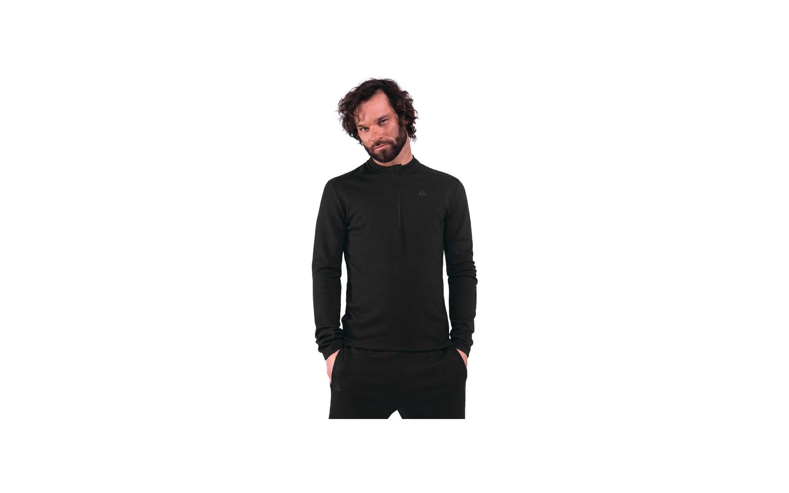 Persona con barba e capelli scuri ricci fino alle spalle, indossa una maglia a maniche lunghe nera e pantaloni neri, mani nelle tasche, sfondo bianco
