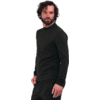 Persona di sesso maschile con capelli scuri e ricci e barba folta, indossa una maglia nera a maniche lunghe e pantaloni neri, in piedi davanti a uno sfondo bianco, leggermente girata di lato, mano sinistra parzialmente in tasca