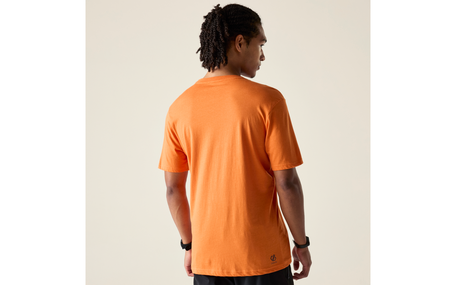 Person von hinten, trägt orangefarbenes T-Shirt und schwarze Shorts, Uhr am Handgelenk, dunkle Dreadlocks, heller einfarbiger Hintergrund