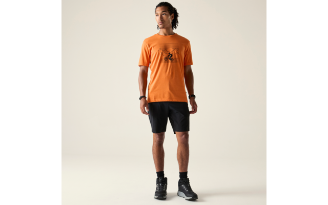Person steht vor hellem Hintergrund, trägt orangefarbenes T‑Shirt mit schwarzer Fahrradgrafik auf der Brust, schwarze knielange Shorts, schwarze Socken und schwarze Sportschuhe; Haare zu Dreadlocks zurückgebunden; Uhr und Armband an den Handgelenken