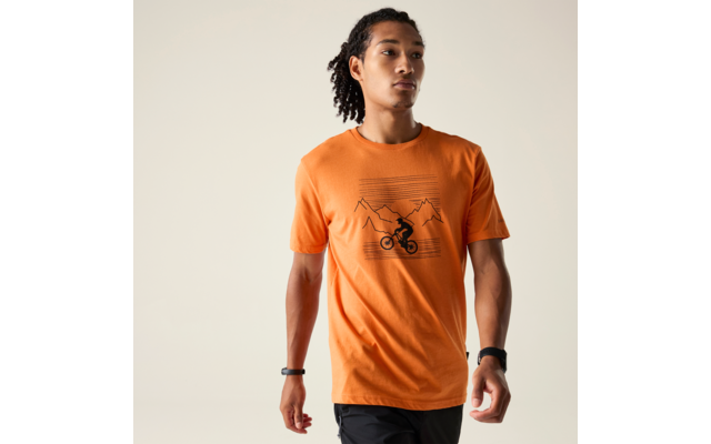 Person mit geflochtenen Haaren trägt orangefarbenes T‑Shirt mit Berg- und Fahrradgrafik, schwarze Shorts, Uhr und Armband an den Handgelenken, Blick nach rechts vor hellem Hintergrund