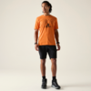 Stehende Person mit zusammengebundenen Dreadlocks, orangenem T‑Shirt mit Fahrradmotiv, schwarzen Shorts und schwarzen Sneakers, schaut nach rechts vor hellem Hintergrund