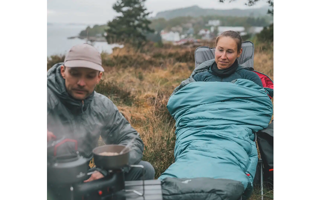 Twee personen zitten buiten, een kookt op een campingkachel