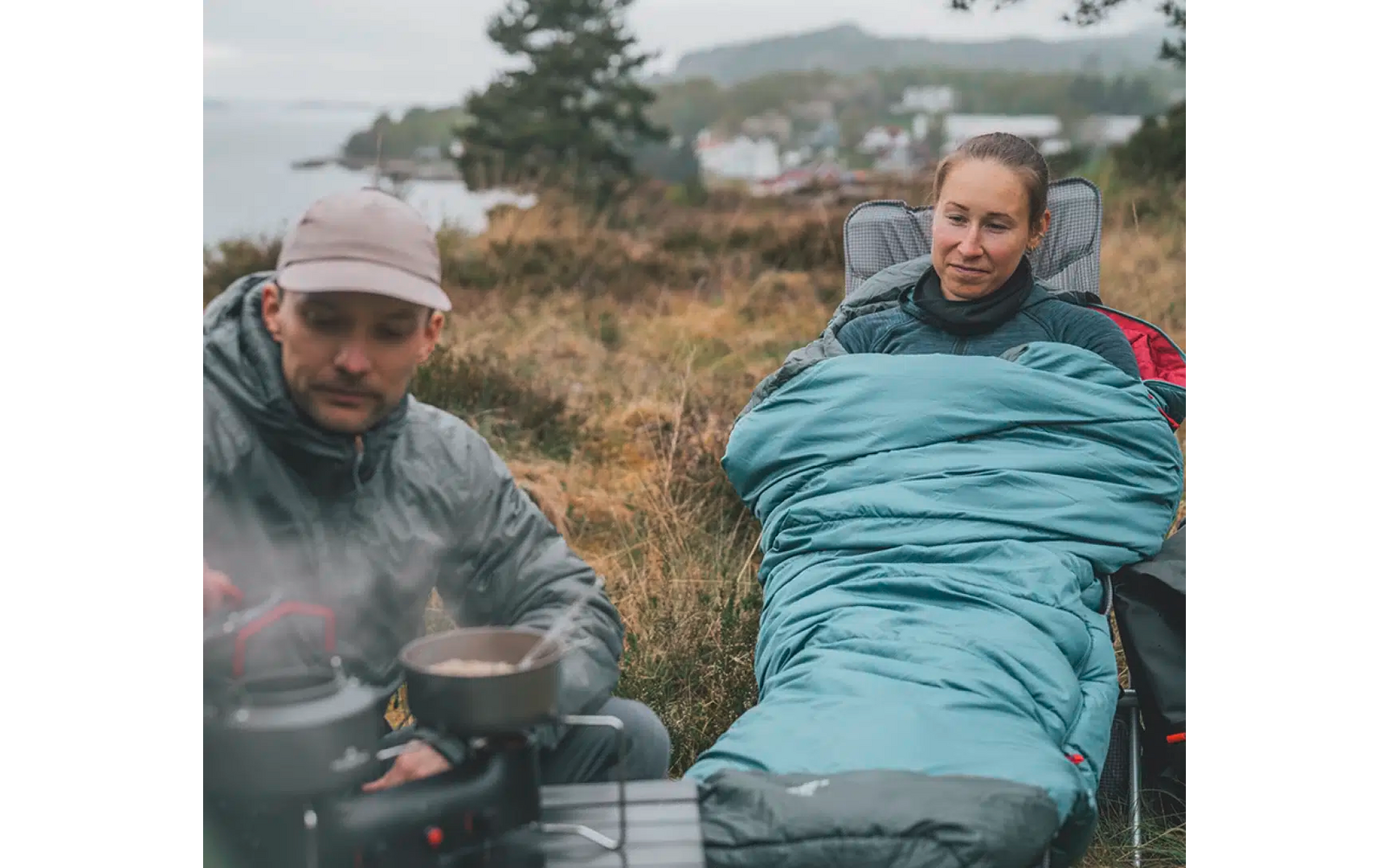 Twee personen zitten buiten, een kookt op een campingkachel