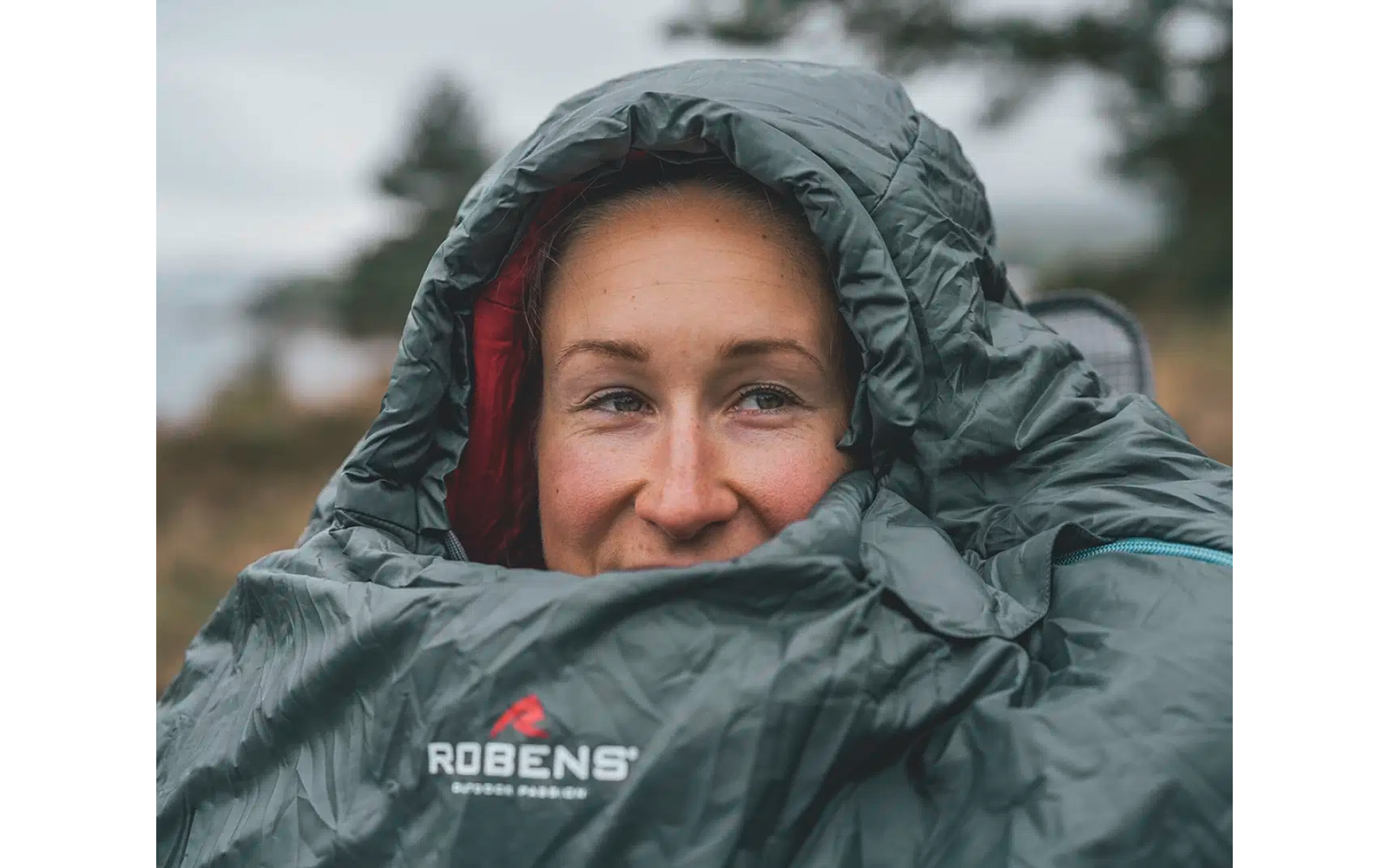 Vrouw draagt een donkergroene outdoor jas met capuchon, kijkt in de camera, vage natuurlijke achtergrond
