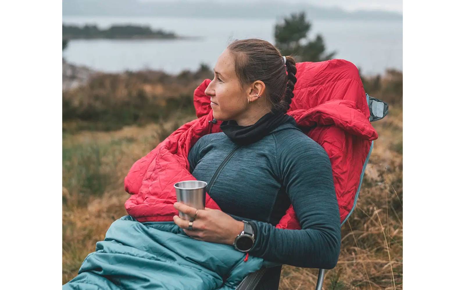 Vrouw zit buiten op gras met een rode rugzak en houdt een kopje in haar hand
