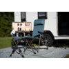Hoge opvouwbare campingstoel, lage opvouwbare kruk en klein opvouwbaar tafeltje met bekers voor een caravan op een patroonmat; wiel en open deur zichtbaar