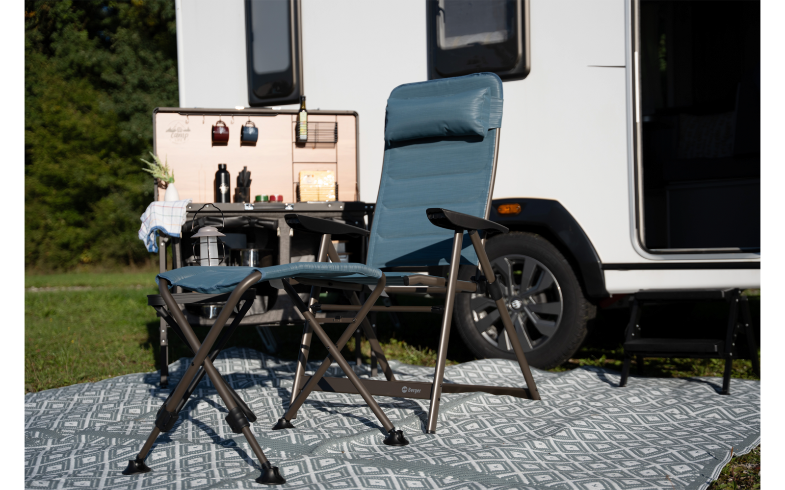 Hoge opvouwbare campingstoel, lage opvouwbare kruk en klein opvouwbaar tafeltje met bekers voor een caravan op een patroonmat; wiel en open deur zichtbaar