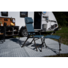 Blauwe campingstoel met hoofdsteun en bijpassende voetenbank op een patroonmat voor een caravan met open deur; links een campingrek met keukengerei, gras op de achtergrond