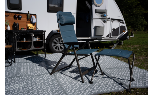 Blauwe campingstoel met hoofdsteun en bijpassende voetenbank op een patroonmat voor een caravan met open deur; links een campingrek met keukengerei, gras op de achtergrond