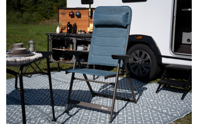 Blauwe vouwstoel op een patroonmat naast een witte camper met geopende zijdeur; links een klein tafeltje met servies; op de achtergrond een open camperkeuken met hangende keukengerei en flessen