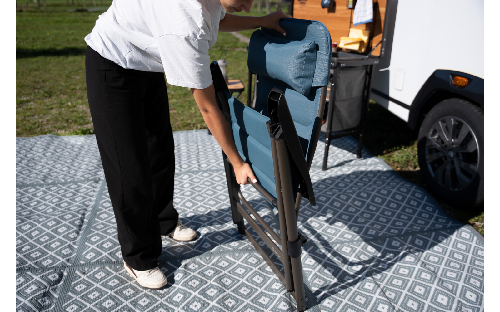 Persoon vouwt een blauwe klapstoel op naast een witte caravan op een grijze buitenmat met patroon