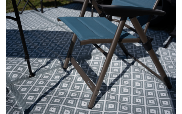 Kleine opvouwbare kruk met blauwe zitting en bruine metalen poten op een grijs-witte mat met geometrisch motief; schaduwen erop