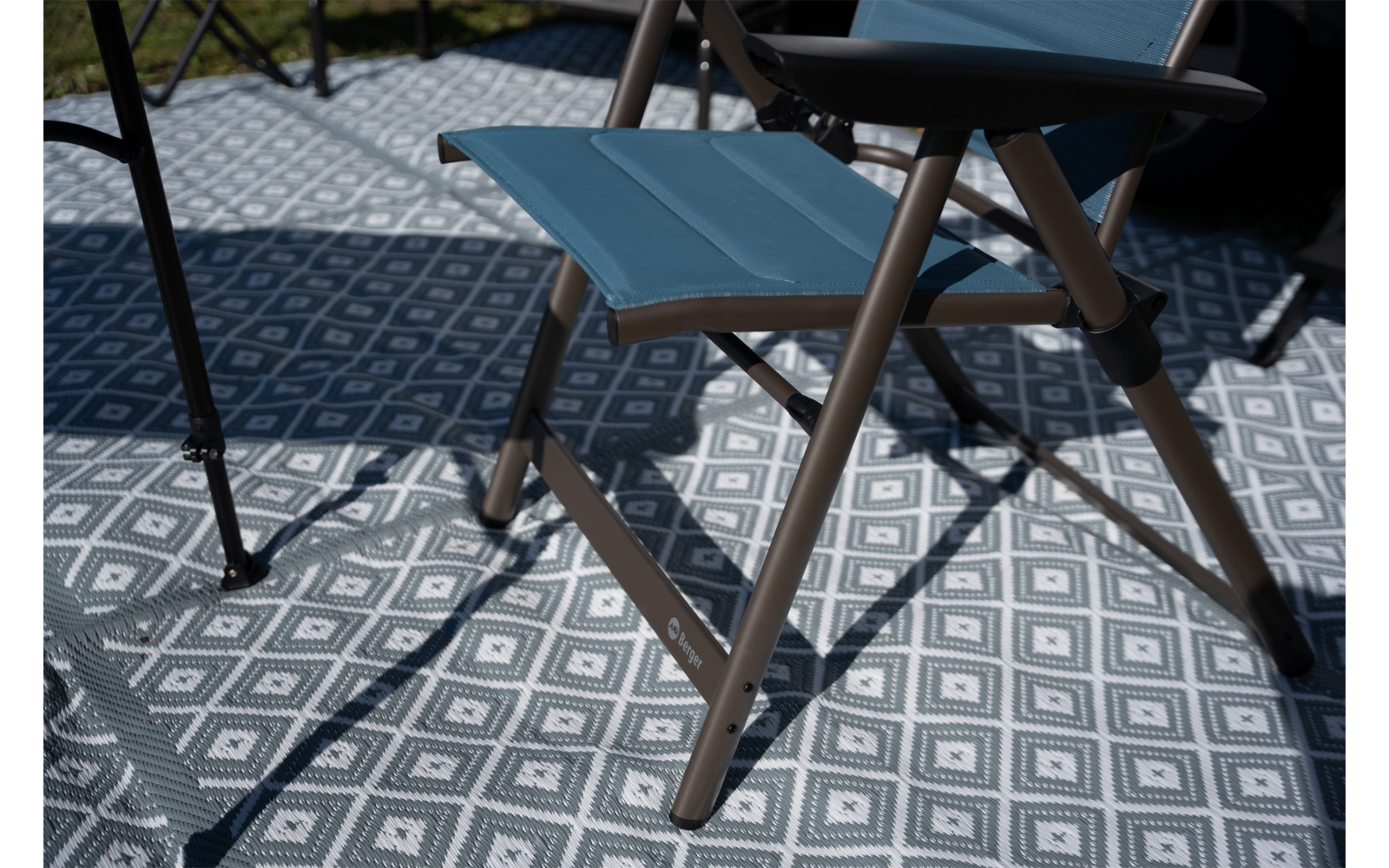 Kleine opvouwbare kruk met blauwe zitting en bruine metalen poten op een grijs-witte mat met geometrisch motief; schaduwen erop