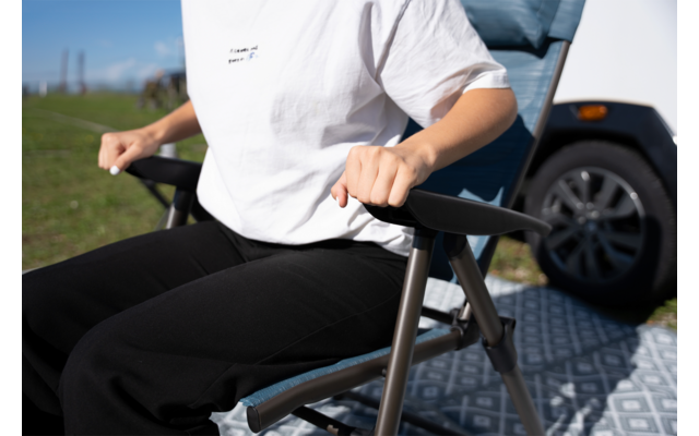 Bovenlichaam van een persoon in wit T‑shirt en zwarte broek, handen op de armleuningen van een blauwe vouwstoel; witte camperband, gras en lucht op de achtergrond