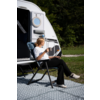 Personne aux cheveux blonds attachés assise sur une chaise pliante devant une caravane blanche, tenant une tasse et un smartphone ; porte de caravane ouverte, herbe, arbres et tapis d'extérieur à motif visibles