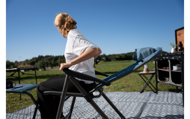 Personne assise sur une chaise pliante, buste tourné vers la gauche, vêtue d'une chemise blanche et d'un pantalon noir, cheveux en chignon ; en extérieur sur un tapis à motifs, zone herbeuse et ciel bleu, table de camping et une autre chaise à droite