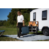 Personne en t-shirt blanc et pantalon noir porte une chaise pliante bleue pliée à côté d’un compartiment extérieur ouvert avec panneau en bois d’une caravane blanche, sur un tapis à motifs sur l’herbe