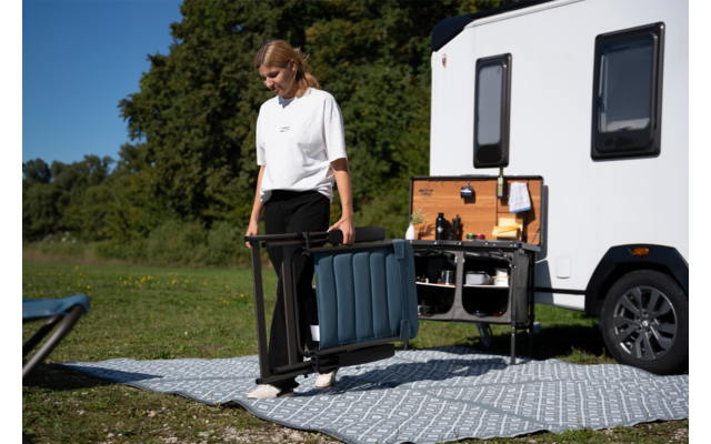 Personne en t-shirt blanc et pantalon noir porte une chaise pliante bleue pliée à côté d’un compartiment extérieur ouvert avec panneau en bois d’une caravane blanche, sur un tapis à motifs sur l’herbe