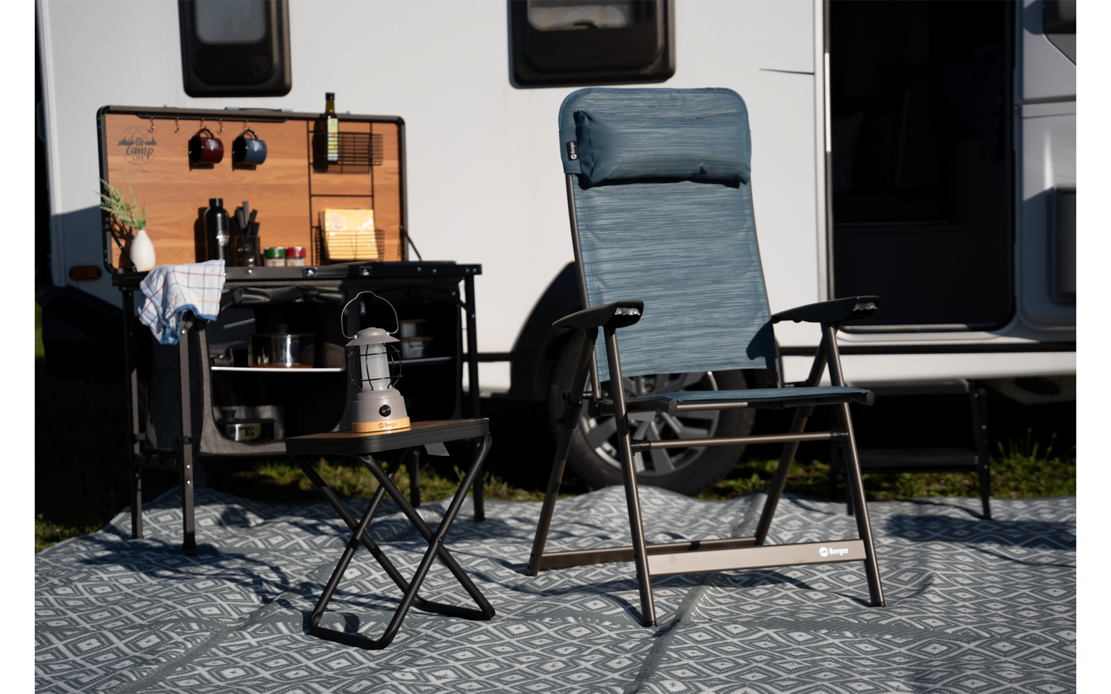 Deux chaises de camping devant une caravane sur un tapis à motifs; petite table pliante avec une lanterne et une tasse; meuble de cuisine de camping à panneau en bois, bouteilles, ustensiles et casserole; porte de la caravane ouverte et fenêtres visibles