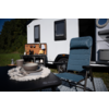 Caravane blanche sur l'herbe, mallette de cuisine de camping ouverte, chaise de camping bleue devant une table avec assiettes, bols et tasses, tapis de sol et arbres en arrière-plan