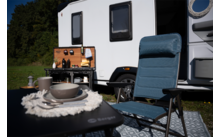Weißer Wohnwagen auf einer Wiese, offene Camping-Küchenbox, blauer Campingstuhl vor einem Tisch mit Tellern, Schalen und Tassen, Bodenmatte und Bäume im Hintergrund