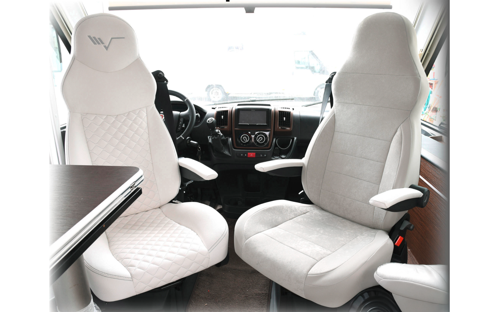 Two white comfortable driver seats in a vehicle with armrests and a table next to the left seat