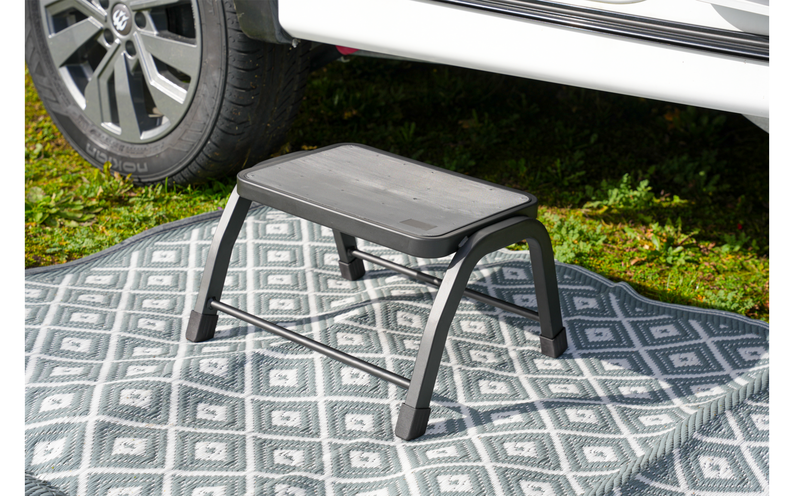 Kleiner schwarzer Kunststofftritt mit vier Beinen auf einem grauen, gemusterten Teppich neben dem Reifen eines weißen Fahrzeugs, Gras im Hintergrund