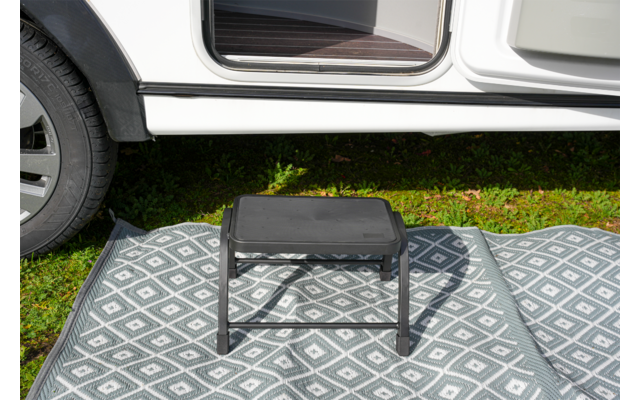 Schwarzer Tritthocker auf grau-weiß gemustertem Außen-Teppich vor offener Tür eines weißen Fahrzeugs, Gras darunter