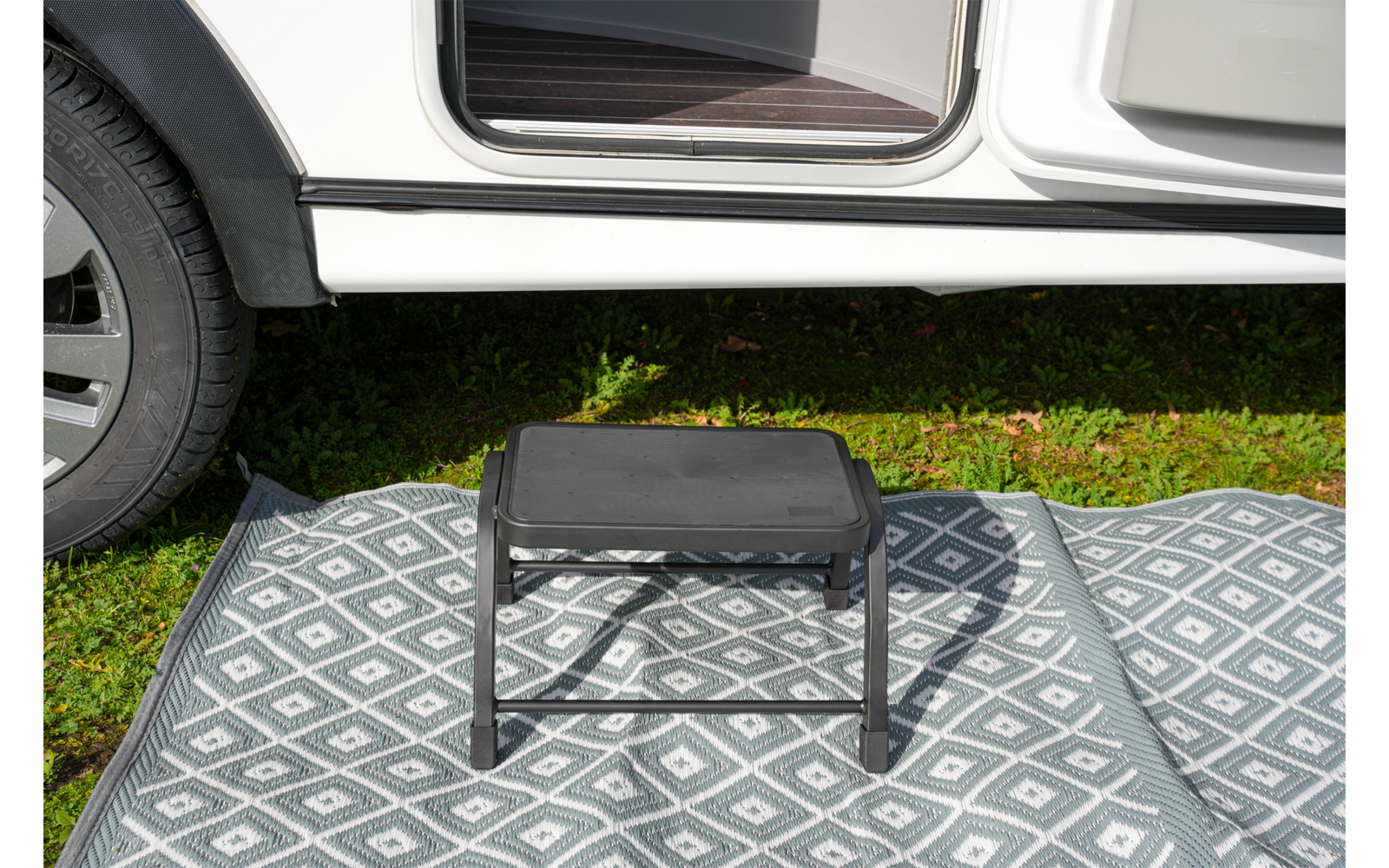 Schwarzer Tritthocker auf grau-weiß gemustertem Außen-Teppich vor offener Tür eines weißen Fahrzeugs, Gras darunter