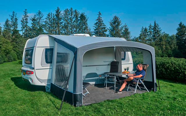 Weißer Wohnwagen mit grauem Vorzelt auf grüner Wiese; Person sitzt auf Klappstuhl unter dem Vordach, kleiner Tisch mit Gegenständen, Nadelbäume und blauer Himmel im Hintergrund