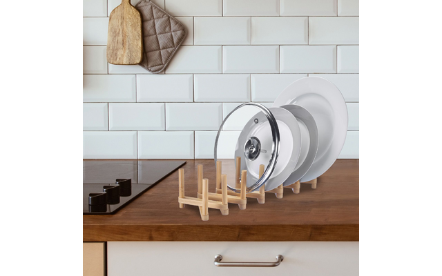 Piano cucina con diversi coperchi di pentole e piatti in un supporto di legno, piastrelle bianche a mattoncino, due taglieri in legno e una presina al muro, piano cottura nero a sinistra