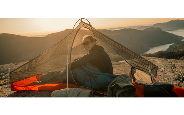 Persoon met zonnebril zit in een tent met gaas op een rotsrichel; bergen, een meer en een oranje lucht zijn zichtbaar op de achtergrond