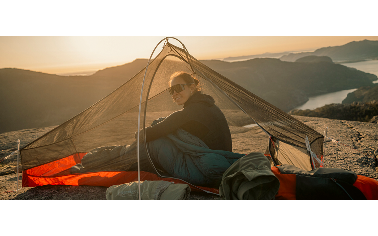 Persoon met zonnebril zit in een tent met gaas op een rotsrichel; bergen, een meer en een oranje lucht zijn zichtbaar op de achtergrond
