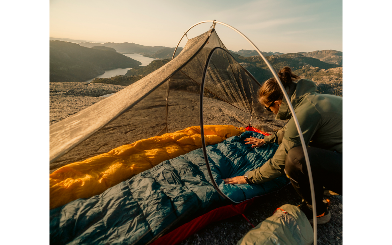Persoon richt een slaapzak in een lichtgewicht tent met muskietengaas op een rotskam, valleien en een meer op de achtergrond