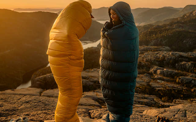 Twee personen staan op een rotsplateau bij zonsondergang, elk in een slaapzakachtig pak (geel, donkerblauw) op matjes; schoenen naast hen; water en bergen op de achtergrond