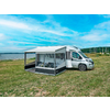 Camping-car blanc avec auvent déployé et véranda amovible sur une pelouse au bord d'une étendue d'eau ; table et chaises visibles dans la véranda ; en arrière-plan eau, rivage, mâts de voiliers et ciel bleu avec des nuages