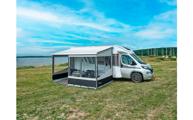 Camping-car blanc avec auvent déployé et véranda amovible sur une pelouse au bord d'une étendue d'eau ; table et chaises visibles dans la véranda ; en arrière-plan eau, rivage, mâts de voiliers et ciel bleu avec des nuages