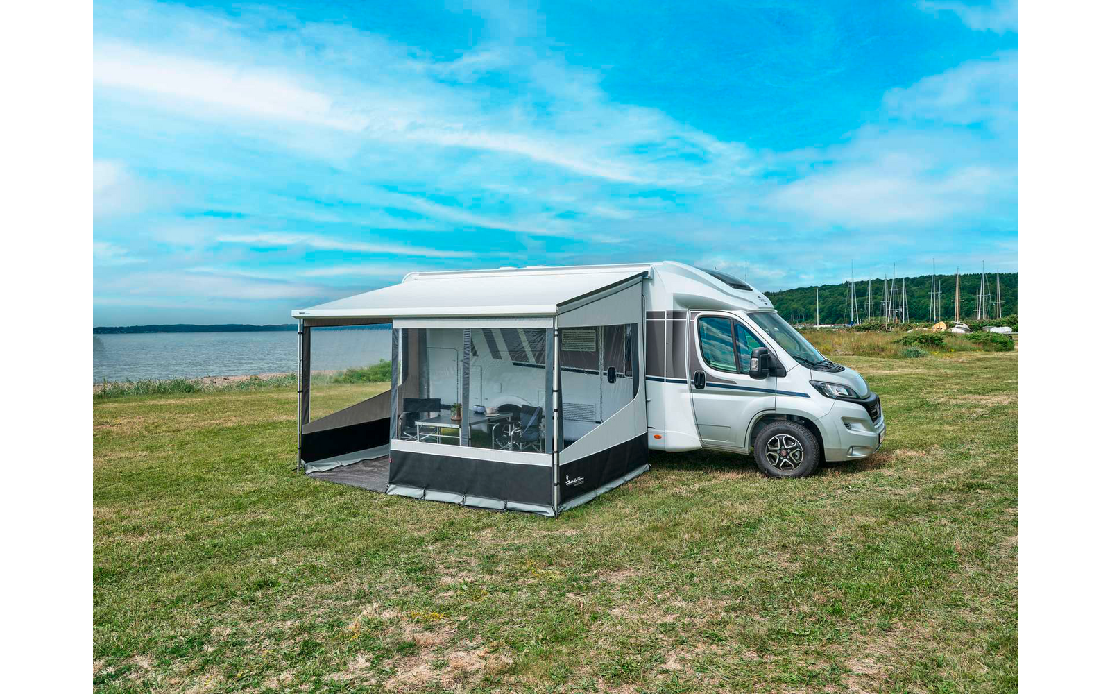 Camping-car blanc avec auvent déployé et véranda amovible sur une pelouse au bord d'une étendue d'eau ; table et chaises visibles dans la véranda ; en arrière-plan eau, rivage, mâts de voiliers et ciel bleu avec des nuages